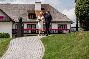 Ein Paar in formeller Kleidung geht Hand in Hand einen gepflasterten Weg entlang und verkörpert die Eleganz einer Hochzeit. Sie stehen vor einem großen, weißen Haus mit Strohdach und blumengeschmückten Balkonen, perfekt für jede Hochzeitsplanung. Die üppige Umgebung erinnert an einen Garten oder eine ländliche Gegend.