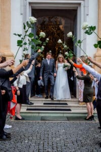 Braut und Bräutigam verlassen ein Gebäude unter einem Torbogen aus weißen Rosen, die liebevoll von einer Gruppe von Menschen gehalten werden. Das weiße Kleid der Braut und der graue Anzug des Bräutigams ergänzen ihre freudige Hochzeit. Der mit kunstvollen Details geschmückte Eingang spiegelt eine sorgfältige Hochzeitsplanung wider.