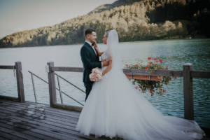 Braut und Bräutigam stehen auf einem Holzsteg an einem ruhigen See, umgeben von üppigen grünen Hügeln. Die Braut trägt ein fließendes weißes Kleid und einen Schleier und hält einen Blumenstrauß in der Hand, während der Bräutigam einen dunklen Anzug trägt. Sie blicken sich liebevoll an und genießen diesen perfekten Moment ihrer Hochzeitsreise.
