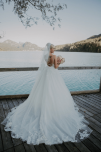 Eine Braut in einem fließenden weißen Kleid und Schleier steht auf einem Holzdeck und hält einen Strauß rosa Blumen in der Hand. Ihr Blick geht auf einen ruhigen See, der von Bergen unter einem klaren Himmel umgeben ist, und sie fängt den perfekten Hochzeitsmoment in ihrer sorgfältigen Hochzeitsplanung ein.