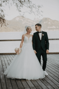 Braut und Bräutigam stehen Hand in Hand auf einem Holzdeck mit Blick auf einen ruhigen See mit Bergen im Hintergrund und fangen die Essenz ihrer Hochzeit ein. Die Braut trägt ein weißes Kleid und hält einen Strauß rosa Blumen, während der Bräutigam, elegant gekleidet in einem dunklen Anzug mit Fliege, sie liebevoll anlächelt.