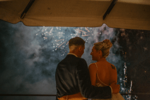 Ein Paar steht umarmt unter einem Baldachin und beobachtet, wie das Feuerwerk den Nachthimmel bei ihrer Hochzeit erhellt. Die Braut trägt ein Spitzenkleid mit einer eleganten Hochsteckfrisur, während der Bräutigam einen dunklen Anzug trägt. Die Szene ist romantisch und festlich.
