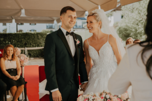 Braut und Bräutigam stehen lächelnd während einer Hochzeitszeremonie im Freien. Die Braut trägt ein weißes Kleid und einen Schleier, während der Bräutigam einen schwarzen Anzug mit Fliege trägt. Sie halten vor einem Geistlichen Händchen und verkörpern damit Monate sorgfältiger Hochzeitsplanung. Im Hintergrund sitzen die Gäste.
