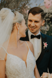 Braut und Bräutigam blicken sich liebevoll an und verkörpern die Essenz einer perfekten Hochzeit. Die Braut trägt ein weißes Kleid mit Spitzendetails und Schleier, während der Bräutigam einen dunklen Anzug mit Fliege und Ansteckblume trägt. Sie stehen inmitten üppiger Vegetation und fangen einen magischen Moment ein.
