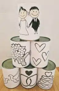 Eine Pyramide aus fünf dekorierten Dosen, geschmückt mit niedlichen Zeichnungen von Braut und Bräutigam, Blumen, Herzen, einer Taube, Eheringen und einem Kuchen auf den Etiketten – alles perfekt für Ihre Hochzeitsplanung. Die Dosen sind ordentlich auf einer Holzoberfläche angeordnet.