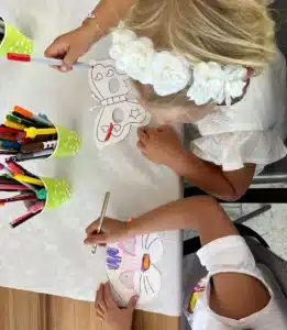 Zwei Kinder sitzen an einem Tisch und bemalen im Rahmen einer spielerischen Hochzeitsfeier Masken mit Markern. Eine Maske ähnelt einem Schmetterling und die andere einer Katze. Der Tisch ist mit einem weißen Tuch bedeckt und von bunten Bechern mit Markern umgeben, was der fröhlichen Atmosphäre der Hochzeit Charme verleiht.