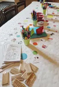 Ein Tisch mit einer weißen Tischdecke ist zum Basteln gedeckt und erinnert an eine kreative Hochzeitsplanungssitzung. Es sind Malbücher, Becher mit Markern, Buntstifte und verstreute bunte Papierausschnitte vorhanden.