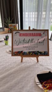 Auf einem Schild auf einer Staffelei steht „KINDERRAUM Wir haben geheiratet Barbara & Leonhard 22. Juli 2023“, mit einem Rand aus Zeichentrickfiguren, perfekt für eine Hochzeit. Es steht auf einem Tisch mit weißer Tischdecke, umgeben von Markern, Buntstiften und Bastelutensilien für Kinder.