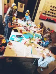 Kinder, die um einen Tisch sitzen und sich mit Kunst- und Bastelaktivitäten beschäftigen, wecken die Kreativität, die einer sorgfältigen Hochzeitsplanung gleichkommt. Der Tisch ist, ähnlich wie bei einer festlichen Hochzeit, mit Zeichenpapier, Buntstiften und Malutensilien bedeckt. Lehrreiche Poster an den Wänden verstärken diese kreative Umgebung.