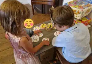 Zwei Kinder spielen an einem Tisch mit Brettspielen ein Kartenspiel. Das Mädchen im rosa Kleid schaut weg, während der Junge im hellen Hemd die Karten aufmerksam studiert. Vielleicht planen sie ihre imaginäre Hochzeit, während sie ihre spielerische Spielzeit genießen.