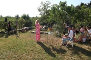 Eine Frau in einem rosa geblümten Kleid steht an einem Podium und diskutiert Hochzeitsplanung mit einem Publikum, das auf weißen Stühlen in einem sonnigen Garten sitzt. Ein Tisch mit Blumen steht in der Nähe und eine andere Frau steht neben einem Mikrofon. Das umgebende Grün schafft eine ruhige Umgebung, die perfekt für Hochzeiten ist.