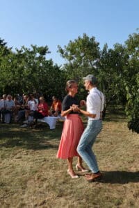 Ein Paar tanzt fröhlich in einer wunderschönen Umgebung im Freien, die an eine romantische Hochzeit erinnert. Umgeben von Bäumen und Gras lächeln die Zuschauer, während die Frau in ihrem rosa Rock herumwirbelt und der Mann selbstbewusst in Jeans und Mütze auftritt und die bezaubernde Essenz von Heiraten einfängt.