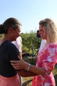 Zwei Frauen lächeln und umarmen sich im Freien unter einem klaren Himmel. Eine hat ihr Haar zurückgebunden und trägt ein schwarzes Oberteil, während die andere, vielleicht in Hochzeitspläne vertieft, lockiges blondes Haar hat und ein rosa-weißes Kleid trägt. Sie scheinen einen Moment der Kameradschaft zu genießen.
