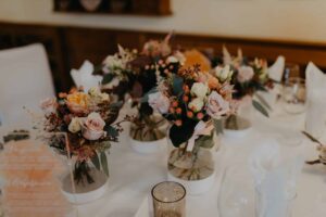 Der Tisch, der sich perfekt für eine Hochzeitsfeier eignet, ist mit sechs exquisiten Blumenarrangements aus rosa Rosen, kleinen orangefarbenen Blüten und üppigem Grün geschmückt. Klare Vasen sind von weißen Servietten, einer Menükarte und einem Votivkerzenhalter aus Glas auf einer strahlend weißen Tischdecke umgeben.