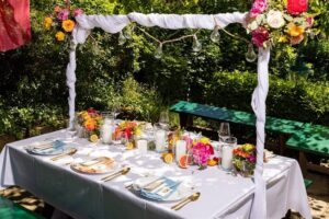 Ein wunderschön dekorierter Tisch im Freien für eine Hochzeitsfeier. Der Tisch ist mit einem weißen Tuch, bunten Blumenarrangements, Glaswaren, Kerzen und Lichterketten ausgestattet. Umgeben von viel Grün ist er perfekt für stilvolle oder intime Hochzeitsfeiern.