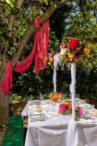 Ein wunderschön gedeckter Tisch im Freien unter einem Baum, geschmückt mit rotem Stoff, bunten Blumen und elegantem Geschirr. Sonnenlicht fällt durch die Blätter und wirft einen warmen Schein auf diese zauberhafte Hochzeitskulisse.