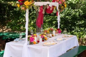 Ein Esstisch im Freien mit weißer Tischdecke, bunten Blumen und Obstdekorationen bildet die perfekte Kulisse für eine Hochzeitsfeier. Darüber hängen Lichterketten und drapierte Stoffe und schaffen das ideale Ambiente für eine Hochzeit inmitten üppigen Grüns.