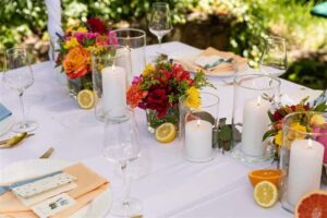 Ein wunderschön gedeckter Tisch im Freien für eine Hochzeit, mit einer weißen Tischdecke, farbenfrohen Blumendekorationen und leuchtenden Orangen und Zitronen. Brennende Kerzen sorgen für eine stimmungsvolle Atmosphäre, während an jedem Gedeck Kristallweingläser mit ordentlich gefalteten pfirsichfarbenen und blauen Servietten stehen – perfekt für Ihre Hochzeitsfeier.