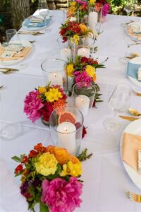 Ein langer Tisch ist elegant für eine Hochzeit gedeckt und mit bunten Sträußen aus rosa, gelben und orangefarbenen Blumen geschmückt. Zwischen den Blumenarrangements sind hohe Glaskerzen eingestreut, die eine festliche und einladende Atmosphäre für diesen besonderen Anlass schaffen.