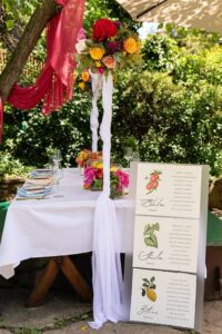 Der wunderschön gedeckte Tisch für eine Hochzeit im Freien besticht durch ein hohes Blumengesteck mit bunten Blüten. Ein weißes Tuch drapiert den Tisch, ergänzt durch eine Menütafel mit eleganten Obstillustrationen und verschiedenen Gerichten. Üppiges Grün umgibt diese perfekte Kulisse für Hochzeiten.