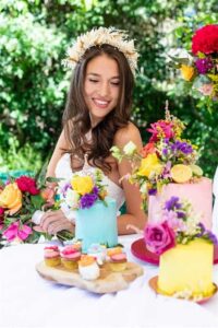 Eine lächelnde Frau mit langen braunen Haaren, einem weißen Kleid und einem Blumenkranz, sitzt im Freien inmitten einer festlichen Szene mit bunten Kuchen und Blumen. Die lebhafte Atmosphäre verleiht dem Tag einen Hauch von Magie, perfekt für eine unvergessliche Hochzeitsfeier.
