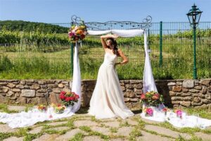 Eine Braut in einem trägerlosen weißen Kleid posiert unter einem mit weißen Vorhängen und bunten Blumen geschmückten Bogen und verkörpert damit elegante Hochzeitsplanung. Die Außenkulisse mit Steinmauer und Grünflächen bildet eine lebendige Kulisse für diese zauberhafte Hochzeitsszene.