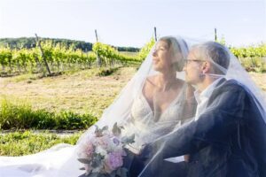Braut und Bräutigam sitzen zusammen unter einem weißen Schleier in einem Weinberg und verkörpern damit die Essenz einer Hochzeit. Die Braut umklammert einen Strauß rosa und weißer Blumen, während beide in die Ferne blicken und über ihre Zukunft inmitten von grünen Reihen nachdenken, die als Leinwand für ihre Hochzeitsplanung dienen.