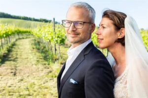 Ein Paar steht in einem Weinberg, die Frau trägt ein Brautkleid und einen Schleier, der Mann einen schwarzen Anzug mit Brille. Im Hintergrund sind grüne Weinreben unter einem klaren blauen Himmel zu sehen, die perfekte Kulisse für ihre Hochzeit. Sie wirken zufrieden und entspannt.