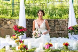 Eine Frau in einem weißen Kleid sitzt lächelnd auf einem weißen Tuch, umgeben von Blumen und brennenden Kerzen vor einer Steinmauer und fängt die Essenz der Hochzeitsplanung ein. Grünes Gras und ein Zaun bilden den Hintergrund, wobei zwei weiße Stoffvorhänge diese zauberhafte Hochzeitsszene elegant umrahmen.