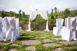 Eine Hochzeitseinrichtung im Freien besteht aus weißen, mit Stoff bezogenen Stühlen, die vor einem dekorativen, mit weißem Stoff behangenen Bogen stehen. Die Kulisse ist von üppigem Grün umgeben und hat im Hintergrund eine Steinmauer. Perfekt für alle, die ihre Traumhochzeit planen.