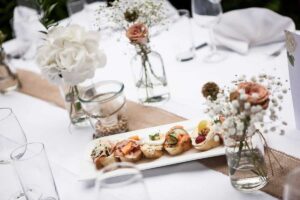 Ein elegant gedeckter Tisch mit weißer Tischdecke und kleinen Sandwiches auf einer weißen Platte als Tafelaufsatz – perfekt für eine Hochzeitsfeier. Daneben stehen Gläser mit weißen und rosa Blumen und leere Weingläser, die eine durchdachte Hochzeitsplanung verkörpern.