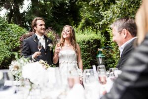 Braut und Bräutigam sitzen lächelnd an ihrem Hochzeitsempfangstisch und erheben ihre Gläser zum Anstoßen. Die Außenanlage ist üppig grün und perfekt für ihre sorgfältig geplante Hochzeitsplanung. Die Gäste sitzen an mit Blumen und Gläsern geschmückten Tischen.