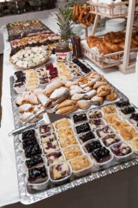 Ein Buffettisch, perfekt für Hochzeitsfeiern, ist übervoll mit Gebäck, Kuchen und Desserts. Tabletts mit Brötchen und Croissants stehen neben einem Glas Nutella und einem Stapel Brezeln. Alles ist elegant auf einer weißen Tischdecke präsentiert und verleiht Ihrer Hochzeitsplanung noch mehr Charme.