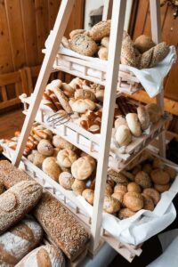 Ein hölzerner Präsentationsständer mit drei Regalen ist vollgestopft mit einer Auswahl an Brot und Brötchen, darunter Baguettes, Brezeln und Brote mit Körnern. Der rustikale Hintergrund schafft eine warme Atmosphäre, die sich perfekt für jede Hochzeitsplanung oder Feier eignet.