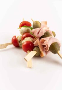 Spieße perfekt für die Hochzeitsplanung: einer mit Kirschtomaten, Mozzarellakugeln und Basilikum, der andere mit Oliven und gefaltetem Schinken, alles elegant angerichtet auf weißem Hintergrund.