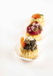 Drei elegante Blätterteighäppchen, in einer Reihe auf einer weißen Oberfläche präsentiert, perfekt für eine Hochzeit. Jedes Häppchen hat einen einzigartigen Belag: eines mit zerkleinertem Grünzeug und Tomaten, ein anderes mit Oliven- und Zwiebelscheiben und das dritte kunstvoll mit einem Aufstrich und einer Garnitur verziert. Ideal für Ihre Hochzeitsplanung.