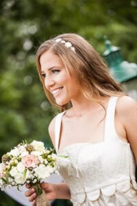 Eine Frau in einem weißen Brautkleid strahlt freudig und umklammert ihren Brautstrauß. Ihr langes Haar, geschmückt mit einem Blumenkopfschmuck, zeugt von der Eleganz einer sorgfältig kuratierten Hochzeitsplanung. Sie steht draußen vor einer Kulisse aus üppigem Grün und fängt die Essenz von Heiraten ein.