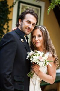 Braut und Bräutigam lächeln in die Kamera und feiern ihren Hochzeitstag. Die Braut hält einen Blumenstrauß in der Hand und trägt ein weißes Kleid, während der Bräutigam einen dunklen Anzug trägt. Sie stehen eng beieinander, wirken glücklich und entspannt in einer Umgebung im Freien und sind dankbar für die reibungslose Hochzeitsplanung.