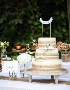 Eine dreistöckige Hochzeitstorte mit rustikalem Design, ideal für Ihre Hochzeit, ist mit weißen Blumen und einem Banner verziert. Sie steht auf einem Holzständer und ist von Gläsern mit Süßigkeiten und Desserts umgeben. Der bezaubernde grüne Blätterhintergrund ergänzt die perfekte Hochzeitsplanungskulisse.