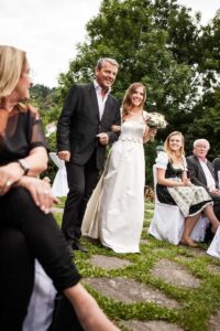 Eine Braut in einem weißen Kleid geht Arm in Arm mit einem Mann im Anzug einen Gang im Freien entlang, umgeben von fröhlichen Gästen. Sie hält einen Blumenstrauß in der Hand, der die Romantik ihrer Hochzeit verkörpert. Üppige grüne Bäume umrahmen diesen wunderschönen Moment der Liebe und Hingabe.