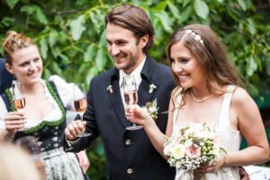 Ein Paar im Hochzeitskleid lächelt, während es bei seiner Hochzeit Champagnergläser hält. Die Braut trägt einen Blumenstrauß und eine Frau in traditioneller Kleidung steht daneben. Grünes Laub dient als Hintergrund und ergänzt perfekt ihre Hochzeitsplanung.