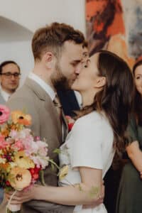 Bei ihrer Hochzeit küsst und umarmt sich ein Paar und hält einen farbenfrohen Blumenstrauß in den Händen. Sie sind von lächelnden Gästen in einem gemütlich dekorierten Raum umgeben und feiern den schönen Moment der Heirat.
