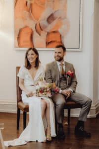 Ein Paar sitzt lächelnd auf Stühlen und feiert seine Hochzeit. Die Frau trägt ein weißes Kleid und hält einen Blumenstrauß, während der Mann einen grauen Anzug mit einer rosa Ansteckblume trägt. Sie sitzen unter einem großen, abstrakten orangefarbenen Gemälde an einer weißen Wand und genießen den Moment ihrer schönen Hochzeit.
