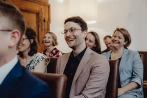 Eine Gruppe von Menschen sitzt in Reihen und lächelt und lacht während einer Hochzeitsveranstaltung. Sie wirken engagiert und glücklich und tragen eine Mischung aus Freizeit- und Geschäftskleidung. Der Hintergrund besteht aus Holzvertäfelungen und weißen Wänden und bietet die perfekte Bühne für unvergessliche Momente bei der Hochzeitsplanung.