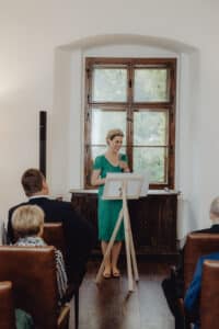 Eine Frau in einem grünen Kleid spricht an einem Podium in ein Mikrofon und diskutiert Hochzeitsplanung. Der Raum, der mit braunen Stühlen und einem Fenster im Hintergrund geschmückt ist, ist mit mehreren sitzenden und aufmerksam zuhörenden Personen gefüllt.