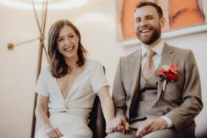 Ein lächelndes Paar sitzt zusammen und hält sich an den Händen. Die Frau trägt ein weißes Kleid und der Mann einen grauen Anzug mit beiger Krawatte und bunter Ansteckblume. Sie strahlen Freude aus, eingefangen in der Wärme ihrer perfekten Hochzeitskulisse.