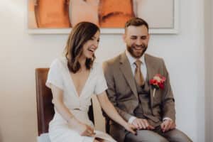 Das vor Freude strahlende Paar sitzt an seinem Hochzeitstag zusammen, lacht und lächelt. Die Frau im weißen Kleid und der Mann im grauen Anzug mit einer roten Ansteckblume teilen diesen glücklichen Moment im Haus, umgeben von abstrakter Kunst.