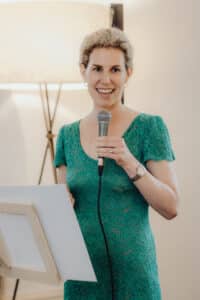 Eine Person mit hellem Haar lächelt, hält ein Mikrofon in einer Hand und steht neben einem Notenständer. Sie bereitet sich möglicherweise auf eine Hochzeitszeremonie vor. Sie trägt ein grünes Spitzenkleid und goldene Creolen. Im Hintergrund steht eine Stehlampe, die zur Atmosphäre der Feier beiträgt.