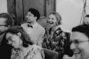 Ein Schwarzweißfoto fängt die Freude einer Hochzeitsfeier ein, bei der die Menschen dicht beieinander sitzen und lachen. In einem formellen Rahmen trägt der Mann Anzug und Fliege, während die Frau in Pailletten glänzt. Die Stimmung ist lebhaft und strahlend.