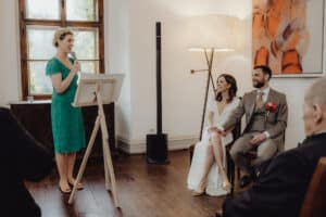 Bei einer wunderschön geplanten Hochzeit hält eine Frau in einem grünen Kleid ein Mikrofon und spricht, während die Braut in Weiß leuchtet und der Bräutigam in seinem grauen Anzug mit einer roten Ansteckblume elegant aussieht. Die Gäste genießen den warm beleuchteten Raum, der bei diesem besonderen Anlass der Hochzeit Freude und Feierlichkeit ausstrahlt.